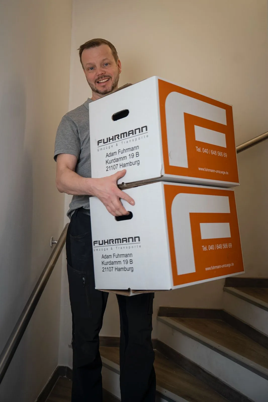 Adam Fuhrmann trägt gebrandete Umzugskartons durch ein enges Treppenhaus