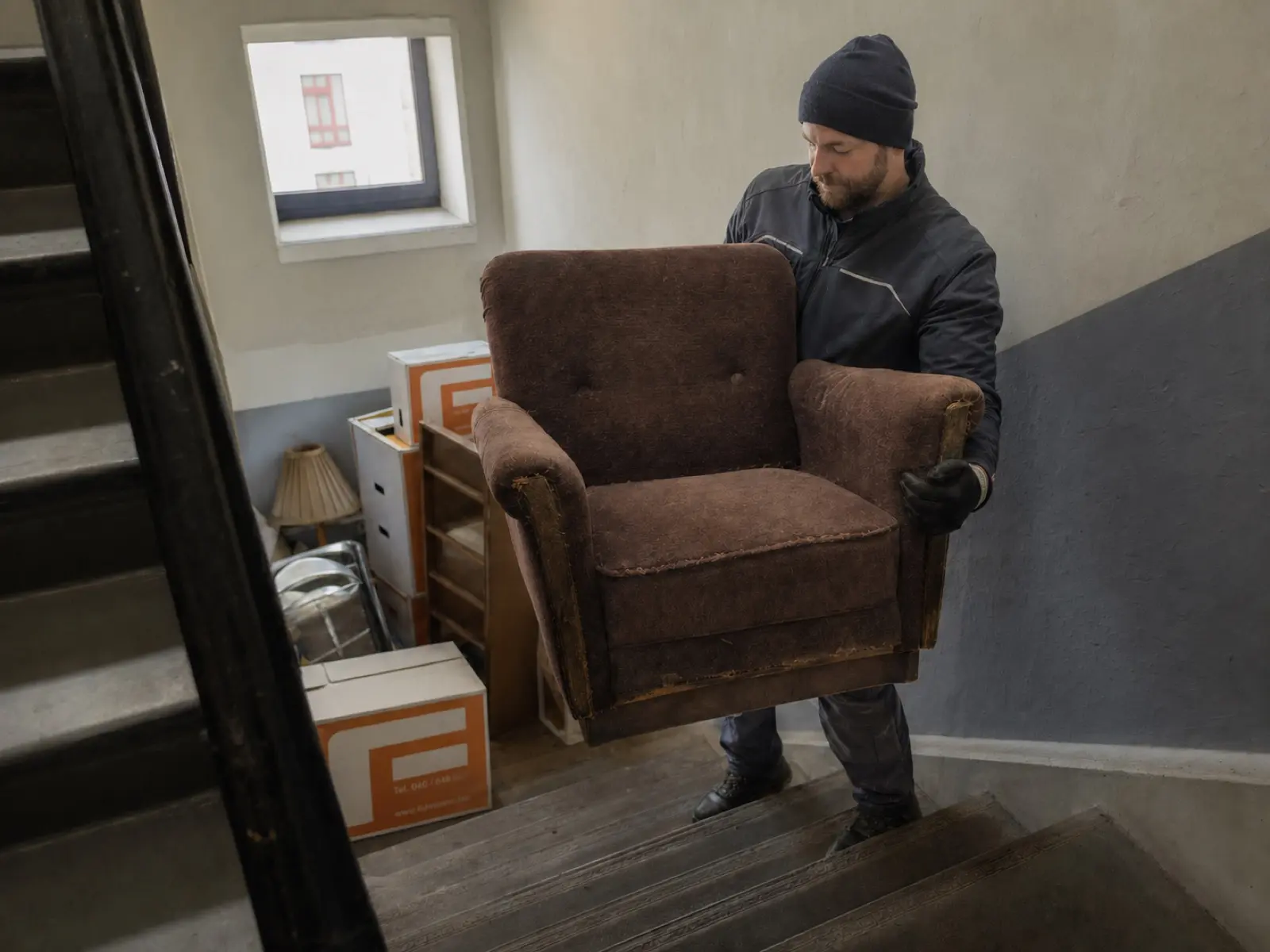 Mover trägt einen alten Sessel ein Treppenhaus hinunter, weitere ausrangierte Möbel im Hintergrund